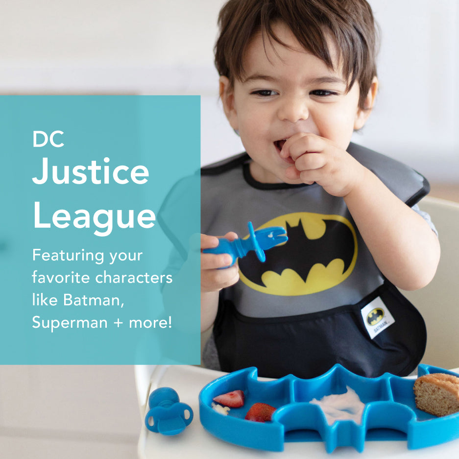 A toddler wearing a Batman-themed bib is seated at a table, eating snacks from a blue Batman-shaped plate. The text on the left reads: DC Justice League Featuring your favorite characters like Batman, Superman + more!.