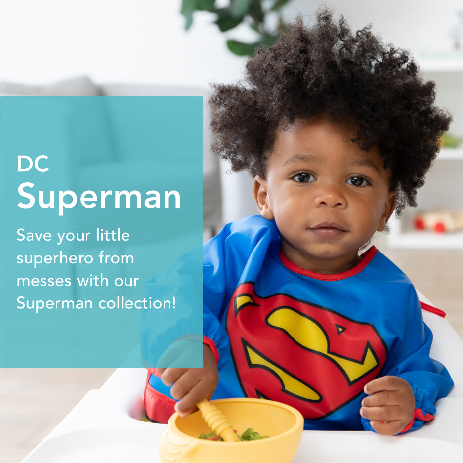 A toddler wearing a Superman bib sits at a table with a bowl of food. Text on the image reads: DC Superman. Save your little superhero from messes with our Superman collection!.