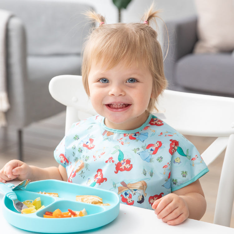 A smiling blonde child with small pigtails wears a blue mermaid patterned bib while eating from a blue divided plate at a white table.