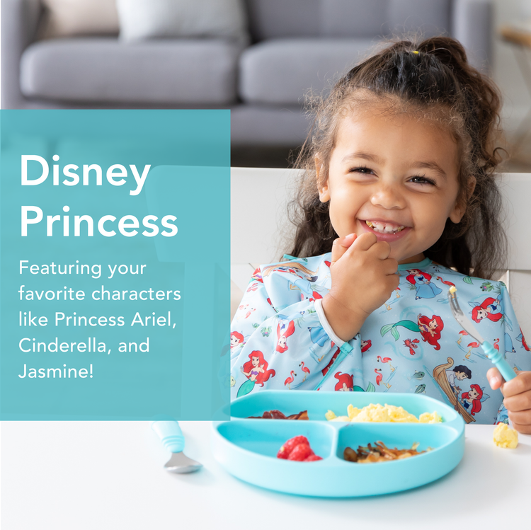 A smiling young girl is sitting at a table eating from a blue divided plate with food. She wears a bib with Disney princesses on it. Text next to her reads, Disney Princess: Featuring your favorite characters like Princess Ariel, Cinderella, and Jasmine!.
