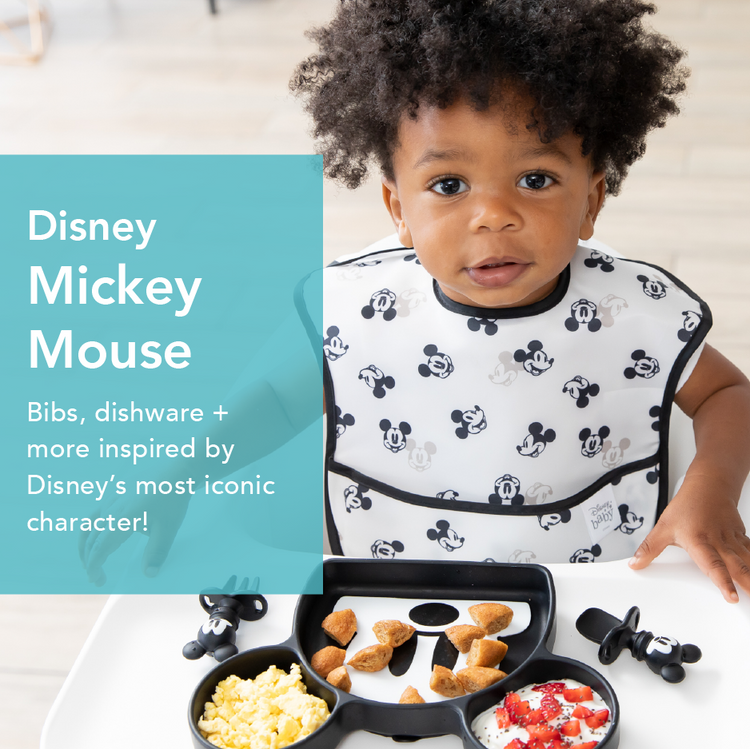 A toddler with curly hair wearing a bib featuring Mickey Mouse faces sits in a high chair. In front of them is a divided plate with scrambled eggs, diced bread, and yogurt with strawberries. Promotional text about Mickey Mouse-themed bibs is visible.