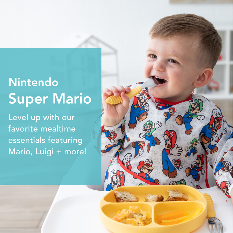 A toddler wearing a bib with Mario designs is eating at a table. The table has a yellow plate with various foods. Text on the image reads: Nintendo Super Mario. Level up with our favorite mealtime essentials featuring Mario, Luigi + more!.