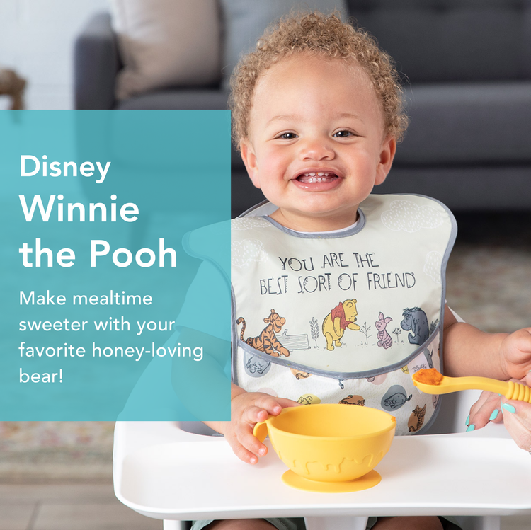 A smiling toddler sitting in a high chair wearing a Winnie the Pooh bib, holding a yellow bowl. An adult hand offers a spoonful of food. The text on the left reads: Disney Winnie the Pooh. Make mealtime sweeter with your favorite honey-loving bear!.