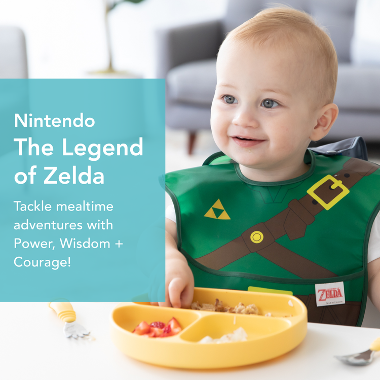 A smiling baby sits at a table wearing a bib inspired by The Legend of Zelda, with a toy sword and shield illustrated on it. The baby is in front of a sectioned yellow plate filled with food. On the left, there is text related to Zelda.