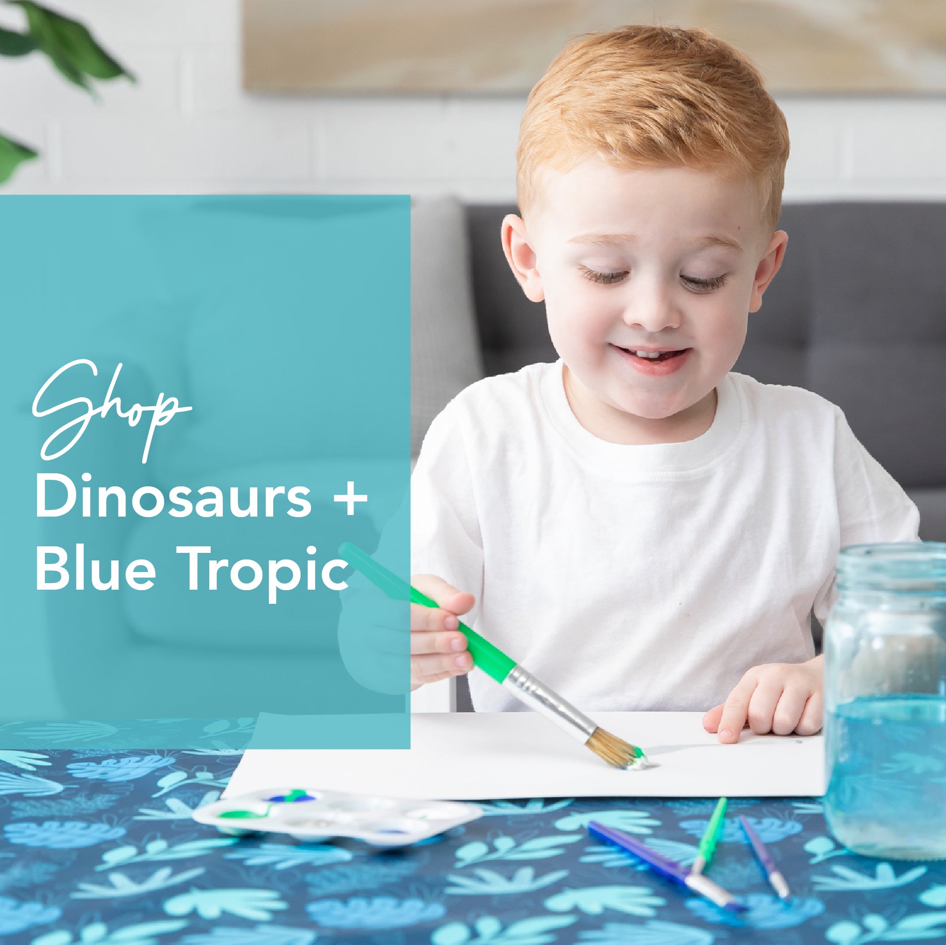 A child with red hair smiles while painting with a green brush at a table covered with a blue dinosaur-patterned cloth. Text on the image says Shop Dinosaurs + Blue Tropic. A jar of water and paintbrushes are on the table.