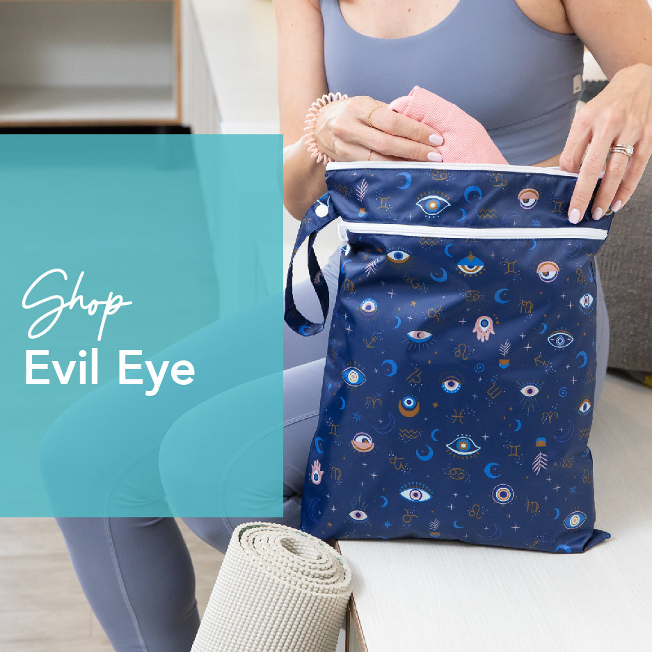 A person in athletic wear places a pink towel into a blue bag adorned with various evil eye designs. The bag is next to a rolled-up yoga mat. Text overlay reads Shop Evil Eye.