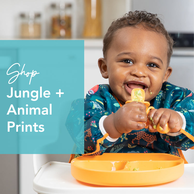 A baby in a high chair wears a jungle-themed bib while eating from an orange plate. The baby smiles and holds a spoon. Text on the image reads Shop Jungle + Animal Prints. The background shows a kitchen.