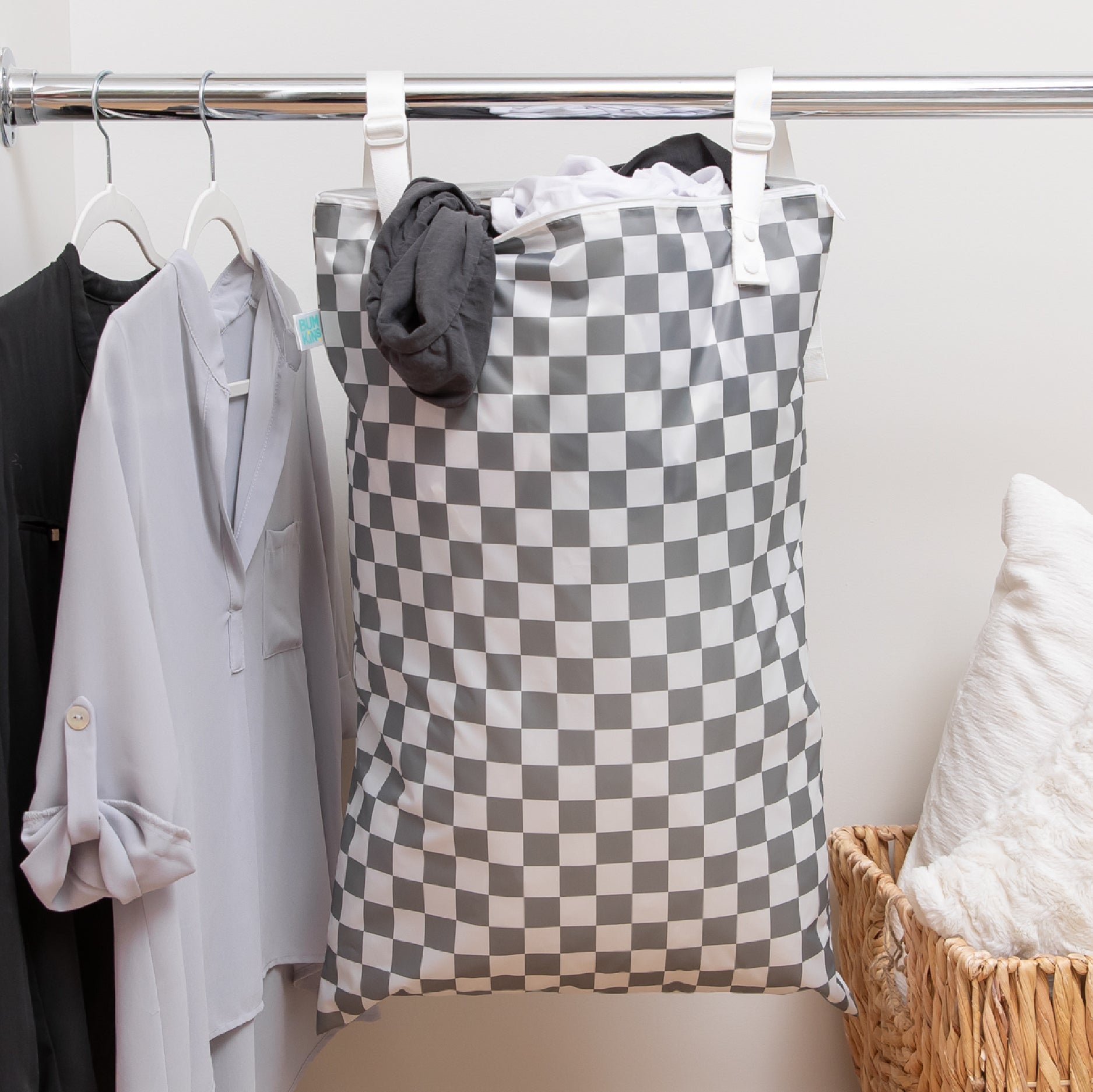 A checkered laundry bag hangs on a rod next to clothes and a basket with pillows.