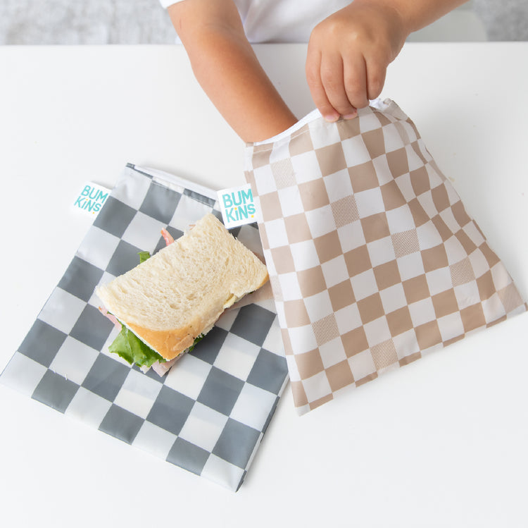 A child places a sandwich into a reusable, checkered Bumkins snack bag on a white table.