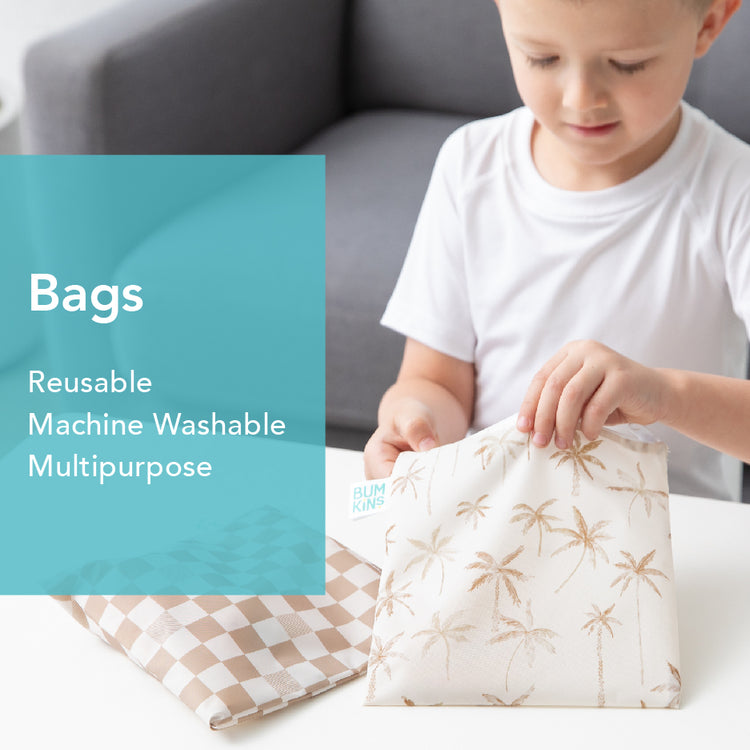 A child in a white shirt sits on a couch, holding a beige reusable bag with palm tree prints. Another checkered bag lies on the table. A teal text box reads: Bags - Reusable, Machine Washable, Multipurpose.