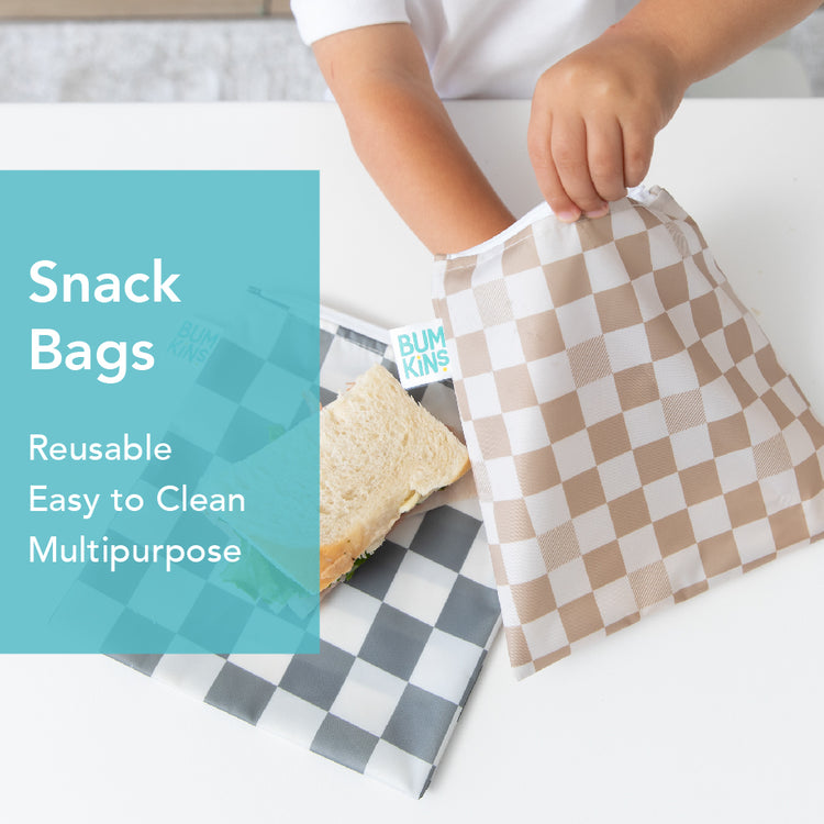 A person places a sandwich into a beige and white checkered reusable snack bag. Another gray and white checkered bag is on a white surface. Text on the left reads: Snack Bags, Reusable, Easy to Clean, Multipurpose.