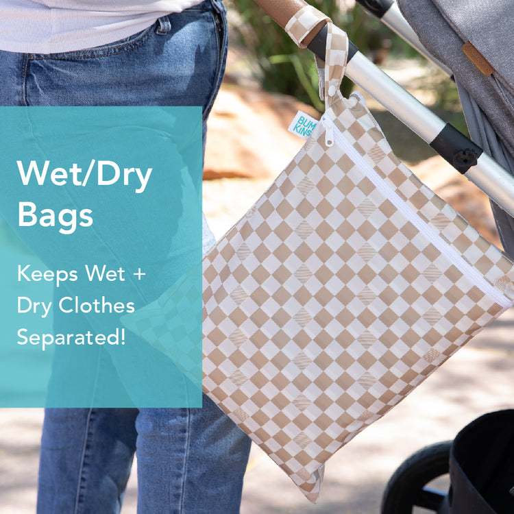 Close-up of a person pushing a stroller with a checkered beige wet/dry bag attached to the handle. The bag has a white zipper and text overlay reads: Wet/Dry Bags. Keeps Wet + Dry Clothes Separated!.