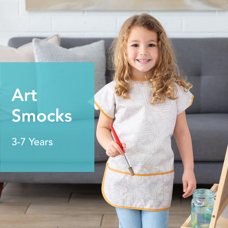 A young child with curly hair stands smiling in a living room, wearing a patterned art smock. They hold a paintbrush next to a painting easel. Text on the left reads Art Smocks 3-7 Years. A couch with cushions is in the background.