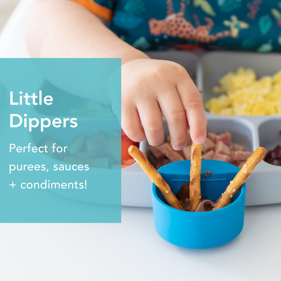 A childs hand dips a stick into a small blue container. The container is next to a tray with scrambled eggs, diced ham, and other foods. Text reads, Little Dippers: Perfect for purees, sauces + condiments!.