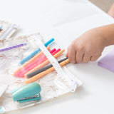 Close-up of a child placing colorful pens in a Bumkins Clear Travel Bag 3 Pack: Palm Daze, with a stapler and sticky notes nearby on a table.
