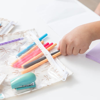 Close-up of a child placing colorful pens in a Bumkins Clear Travel Bag 3 Pack: Palm Daze, with a stapler and sticky notes nearby on a table.