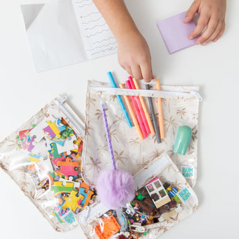 A child's hand reaches for colored pens from Bumkins Clear Travel Bag 3 Pack: Palm Daze filled with toys and art supplies at a white table.