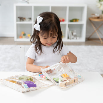 A young girl examines toys in a Bumkins Clear Travel Bag 3 Pack: Palm Daze at a white table in a bright room, wearing a white bow.