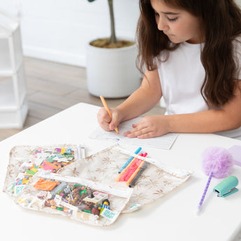 A girl draws at a white table with colored markers and a fluffy pen, near Bumkins Clear Travel Bag 3 Pack: Palm Daze filled with toy blocks.