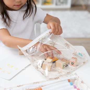 A child places wooden blocks into a Bumkins Clear Travel Bag 3 Pack: Palm Daze, with paper and markers nearby on a white table.