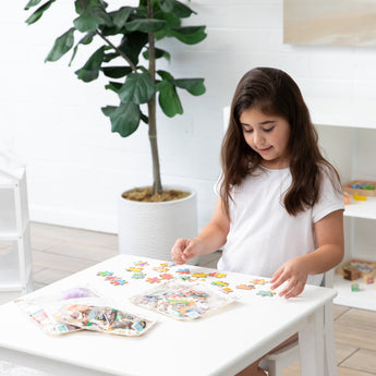 A girl in a white shirt assembles a colorful puzzle at a table, surrounded by Bumkins Clear Travel Bag 3 Pack: Palm Daze and a potted plant.