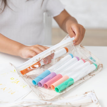 A child organizing colorful markers in a Bumkins Clear Travel Bag 3 Pack: Palm Daze on a white table.
