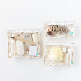 Bumkins Clear Travel Bag 3 Pack: Palm Daze holds toiletries like cotton pads, shampoo, a hairbrush, and a clip on a white background.