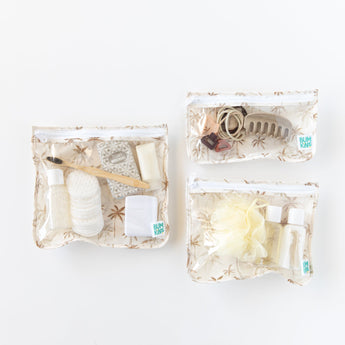Bumkins Clear Travel Bag 3 Pack: Palm Daze holds toiletries like cotton pads, shampoo, a hairbrush, and a clip on a white background.