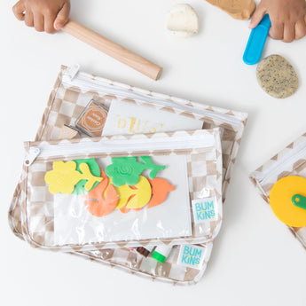 A child plays with colorful tools and clay next to Bumkins Clear Travel Bag 3 Pack: Sand Check filled with art supplies on a white table.