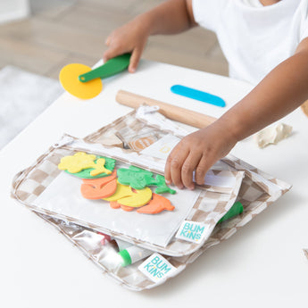 A child plays with a toy pizza cutter and play dough as Bumkins Clear Travel Bag 3 Pack: Sand Check with art supplies sits nearby on a table.