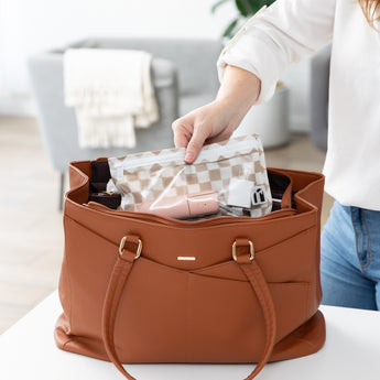 A person places a Roamio by Bumkins Clear Travel Bag 3 Pack: Sand Check with electronics in a brown purse on a white table.