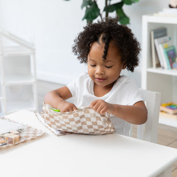 A curly-haired child eagerly unzips a Bumkins Clear Travel Bag 3 Pack: Sand Check, surrounded by books and a plant at a white table.