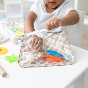 A child in a white shirt reaches for modeling clay from a Bumkins Clear Travel Bag 3 Pack: Sand Check at a white table with art supplies.