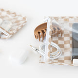 Close-up of a Bumkins Clear Travel Bag 3 Pack: Sand Check with a hair clip and coiled charging cable on a white background near an earbud case.