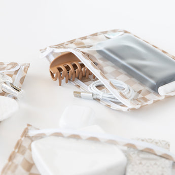 Close-up of Bumkins Clear Travel Bag 3 Pack: Sand Check holding hair clips, USB cables, and personal items on a white background.