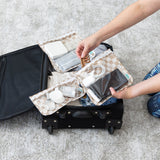 A person packing a suitcase with Bumkins Clear Travel Bag 3 Pack: Sand Check on a gray carpet, holding travel essentials and electronics.