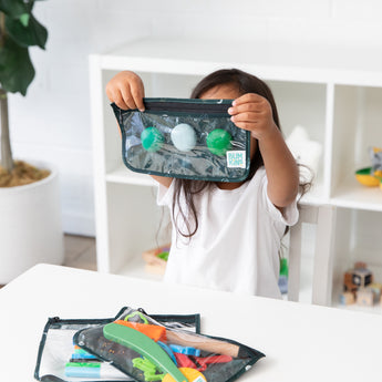 A child inspects items in a Bumkins Clear Travel Bag 3 Pack: Starry Cactus at a white table, while a plant adds a natural touch.