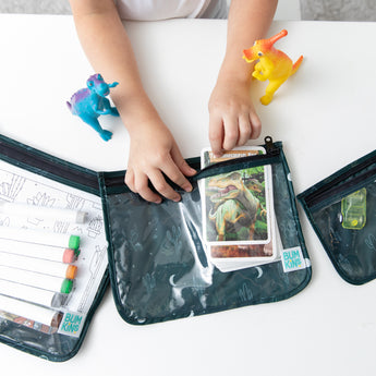 A child puts a card into a Bumkins Clear Travel Bag 3 Pack: Starry Cactus, surrounded by dinosaur toys and art supplies on a white table.