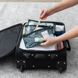 A person places toiletries into a Bumkins Clear Travel Bag 3 Pack: Starry Cactus in an open suitcase on a gray carpet.