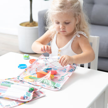 A young girl with long blonde hair places colorful toys into a Bumkins Clear Travel Bag 3 Pack: Boots at a white table.
