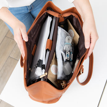 A person opens a brown purse on a white table, revealing notebooks and a Roamio by Bumkins Clear Travel Bag 3 Pack: Disco Check inside.