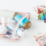Close-up of Bumkins Clear Travel Bag 3 Pack: Yoga Pals holding travel-sized bottles, sunglasses, and hair accessories on a white background.