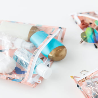 Close-up of Bumkins Clear Travel Bag 3 Pack: Yoga Pals holding travel-sized bottles, sunglasses, and hair accessories on a white background.