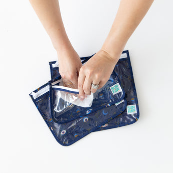 A person uses a wipe to clean the inside of Bumkins Clear Travel Bag 3 Pack: Evil Eye on a white table.