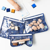 A child's hand plays with stamps beside Bumkins Clear Travel Bag 3 Pack: Evil Eye, filled with wooden toys on a white table.