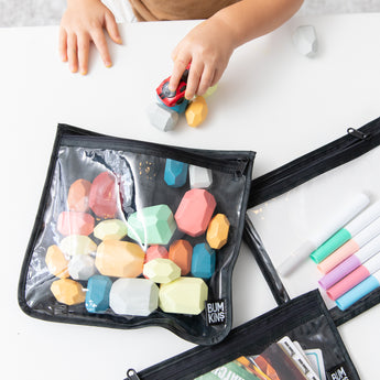 A child plays with a toy car at a white table while Bumkins Clear Travel Bag 3 Pack: Rich Black hold blocks and markers nearby.