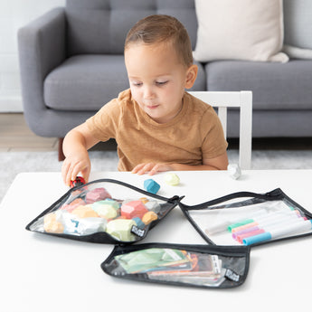A young child plays as Bumkins Wet Bag + Clear Travel Bag 3 Pack Set keeps blocks and markers organized at a white table.