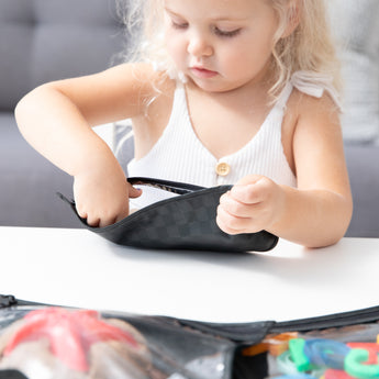 A blonde girl reaches into a Bumkins Clear Travel Bag 3 Pack: Rich Black at a white table.