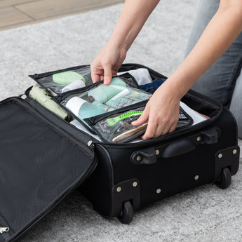 A person packs bags from Bumkins The Chaos Coordinator Deluxe 5-Piece Set: Rich Black filled with toiletries into a suitcase on a gray rug.