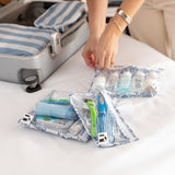 A person packs toiletries into Roamio by Bumkins Clear Travel Bag 3 Pack: Shells on a white bed near an open gray suitcase.